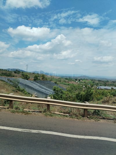 The image shows a roadside view in what appears to be a rural area of Thôn 4, Vietnam. The foreground features an asphalt road with a white lane marking and a metal guardrail. Beyond the guardrail, the terrain slopes downward, revealing a landscape of green vegetation, including trees and crops. A section of the hillside is covered with dark netting. In the distance, a series of wind turbines are visible against a backdrop of hazy mountains and a partly cloudy sky. An electrical transmission tower can also be seen on the ridge.