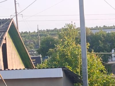 The image depicts a view of residential rooftops and trees against a bright sky. In the foreground, the weathered wooden side of an A-frame structure with a green, corrugated roof is visible. To the right, a white, ribbed soffit runs along the edge of a lower roof. A grey pole stands prominently in the mid-ground, obscuring parts of the scene. Beyond these structures, a dense canopy of green trees with some yellowing leaves fills the mid-ground, interspersed with hints of rooftops and what appear to be residential buildings nestled on a gently sloping hillside. Power lines crisscross the sky in the background, supported by a utility pole. The overall setting suggests a rural or suburban environment, possibly within Piatra Albă, Moldova, given the typical architectural styles and vegetation observed. There are no people or animals visible in the frame. The lighting indicates it is daytime, with the sun high in the sky.