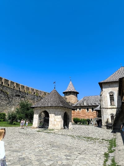 The image displays the interior courtyard of the Khotyn Fortress in Khotyn, Ukraine, observed under a clear, cloudless blue sky. The ground is paved with irregular grey and beige stones, with some green grass growing between the cracks, particularly along the right side.

In the mid-ground, a light-colored stone pavilion with a dark, multi-faceted conical roof stands centrally. This structure features multiple arched openings, at least two of which are visible and contain dark metal grates or gates. A small finial or flag is mounted at the apex of its roof.

To the left, a tall, thick stone fortress wall, dark grey and rugged, extends from the mid-ground towards the background, supporting some sparse green vegetation. In the background, behind the pavilion, a prominent cylindrical stone tower with a dark, pointed conical roof is visible, forming part of the main fortress structure.

On the right side of the image, a series of stone buildings with grey tiled roofs are present. The closest building on the immediate right features a distinctive arched window with dark framing and light trim. Further back and to the right, another building with reddish-brown walls and smaller, rectangular openings can be seen. Wooden steps or a raised platform are visible near the building on the far right.

Three individuals are present in the courtyard. In the lower-left foreground, partially visible from behind, is a person with blonde hair wearing a light-colored top. In the mid-ground, to the left of the central pavilion, two individuals, likely females, are walking away from the camera. One wears a patterned light-colored dress, and the other wears shorts and a light top. The scene is illuminated by strong natural sunlight, indicating daytime.