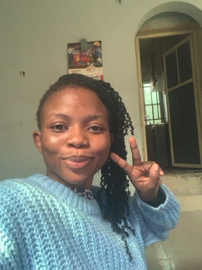 This is an indoor photograph of a young adult female, taken in Onibudo, Nigeria. The subject, positioned centrally, has a dark complexion, dark eyes, and dark braided hair that partially falls over her left shoulder. She faces the camera with a faint smile, and her left hand is raised beside her face, with the index and middle fingers extended to form a 'V' gesture (peace sign). She wears a light blue, textured, long-sleeved sweater over what appears to be a patterned garment visible at the neckline. The background features a plain light-colored wall. A calendar or poster, displaying market items and the word 'ENGAGEMENT', is clipped to the wall above the subject's head. To her right, an open arched doorway reveals an interior space with a window featuring dark grilles, through which a soft green light is visible.