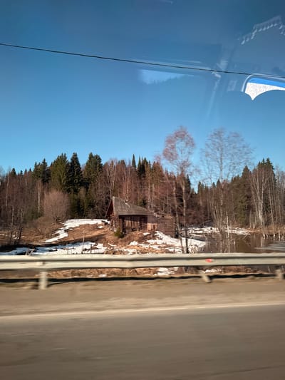 This image, captured from within a vehicle, presents a serene rural landscape, likely in Okulovka, Russia, during late winter or early spring. The perspective is from a window looking outwards.
On the left side of the frame, reflections on the window glass reveal elements of the vehicle's interior, including a light blue surface, possibly part of the ceiling or a panel, and a decorative white, textured object, perhaps a headrest or curtain. Faint reflections of bare trees and the bright blue sky are also superimposed on this area, along with a thin, dark vertical line that might be an antenna or wire.
The central and dominant part of the image showcases the external environment under a clear, vibrant blue sky. A dense forest line dominates the midground, featuring a mix of tall evergreen trees and numerous deciduous trees, many of which are still bare, some displaying a reddish-brown tint on their branches. Tucked among these trees, a rustic wooden house with a dark, sloping roof is visible, suggesting traditional architecture. The ground beneath the trees and around the house is covered in scattered patches of melting snow, interspersed with brown earth and dried vegetation, indicative of a transitional season.
To the right, a multi-lane asphalt road or highway runs parallel to the forest. A grey metal guardrail separates the wooded embankment, with its snow patches, from the roadway. The road surface appears clear, with a faint white line marking its edge on the far right. The bright, clear lighting suggests it is daytime, possibly late morning or early afternoon, with sunny and cold weather. There are no people or discernible text in the image.