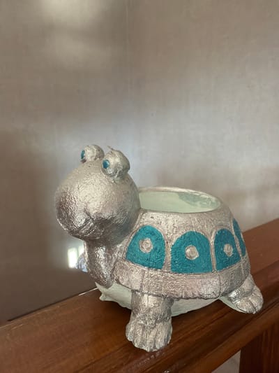 A silver, turtle-shaped planter with teal accents sits on a wooden surface. The planter is facing to the left of the frame, with its head and front legs visible. The background is a textured, beige wall. The wooden surface appears to be a shelf or railing. The lighting in the image suggests it is indoors, possibly in a home or shop in İzmit, Turkey, though no specific landmarks are visible.