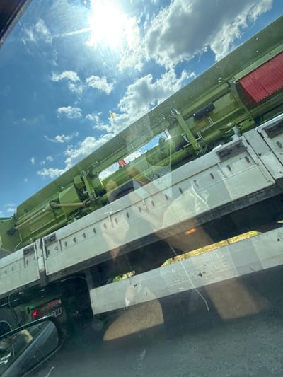 This media file captures a reflection in what appears to be a vehicle's window, oriented sideways. The reflection shows a bright, sunny sky filled with fluffy white clouds, indicating a clear day. The sun is a strong, bright source of light, creating lens flare and highlighting the texture of the clouds.
The main subject of the reflection is the side of a large, industrial vehicle, likely a truck or trailer. It has a long, segmented body, primarily painted in a pale, off-white color. Below this main body, there is a complex green metal structure, possibly part of the vehicle's undercarriage or a piece of machinery. This green section is detailed with various pipes, hoses, and mechanical components.
In the distance, through the reflection, a hint of a landscape with greenery and sunlight can be seen. The foreground of the reflection shows a dark, possibly road surface, with reflections of streetlights or vehicle lights visible as orange and red glows.
There are some notable details within the reflection: a small yellow object, possibly a flag or marker, is visible against the blue sky. On the white segmented part of the vehicle, there are regularly spaced dark markings, possibly hinges or latches. The overall impression is of a daytime scene, likely afternoon given the bright sunlight. The perspective suggests the viewer is inside another vehicle, looking out and seeing the reflection of the sky and the vehicle beside them. No people are clearly discernible in the reflection. There is a license plate visible on a vehicle in the distance within the reflection that reads "2 MA".