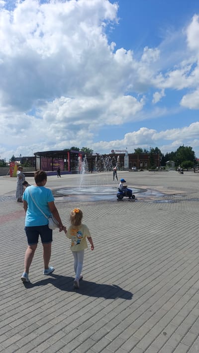 This is a vertical, outdoor, daytime photograph taken on a sunny day with scattered clouds. The scene is a paved plaza with a fountain in the center. In the foreground, a woman in a blue t-shirt and denim shorts walks hand-in-hand with a young girl wearing a yellow t-shirt with a cartoon duck on it and white leggings. They are walking away from the camera, casting long shadows on the brick-like pavement.

In the middle ground, a child wearing a blue cap rides a small blue toy vehicle on the wet pavement around the fountain. The fountain is active, with water spraying upwards. Beyond the fountain, a stage structure with red accents is visible. Further in the background, there are buildings, including one with a sign that reads "Пятёрочка" (Pyaterochka), a Russian supermarket chain. Several other people are scattered throughout the plaza, some walking, and one person appears to be near the stage. The sky is blue with large, fluffy white clouds, indicating fair weather. The overall mood appears to be relaxed and leisurely, suggesting a public space used for recreation. The visible text "Пятёрочка" and "МОЛОДЕЖНАЯ" on the stage structure provide location cues.