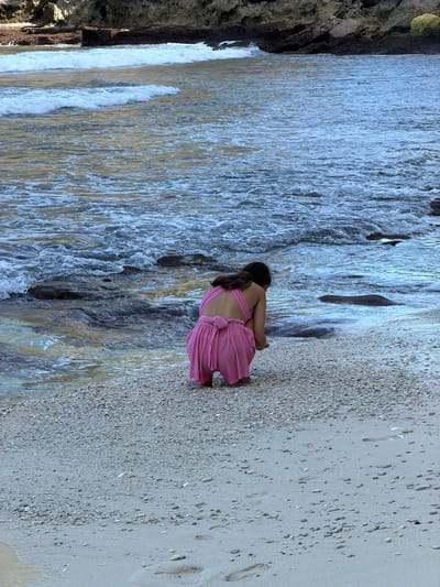 A young girl in a pink dress is crouching on the beach in Pejukutan, Indonesia. She is near the water's edge where the water meets the sand. The beach is composed of a mix of sand and small pebbles. To the left of the image, there is a rocky formation. The overall color palette is a mix of blues, browns, and pinks.