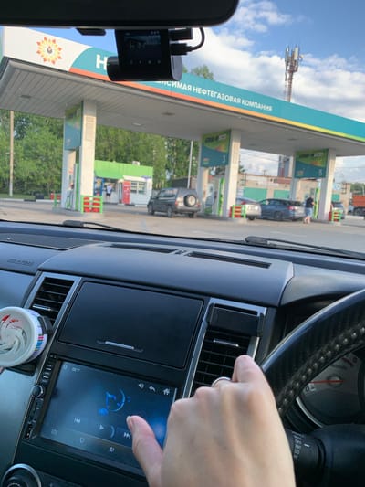 The image shows a view from inside a car, looking out at a gas station. The car's dashboard and steering wheel are visible in the foreground, with a hand reaching towards the car's infotainment system. The infotainment screen displays a music player interface with various controls. A dashcam is mounted on the windshield to the left.
Outside the car, the gas station appears to be operational, with pumps visible. There are several cars at the gas station, including a dark-colored SUV and a lighter-colored car. People are also visible near the pumps. The gas station's canopy is labeled with Cyrillic text, which appears to be "ЛУИСИМАЯ НЕФТЕГАЗОВАЯ КОМПАНИЯ" (LUISIMAYA NEFTEGAZOVAYA KOMPANIYA), likely a company name. There are also numbers "3" and "5" on green signs, possibly indicating pump numbers or fuel types.
In the background, a cell tower can be seen against a sky with scattered clouds. The overall impression is a typical scene at a gas station on a bright day, possibly during the daytime given the lighting. The scene is set in Belogorsk, Russia, as indicated by the context. There are no strong indications of emotion, but the presence of a hand interacting with the car's system suggests a person is driving or a passenger.