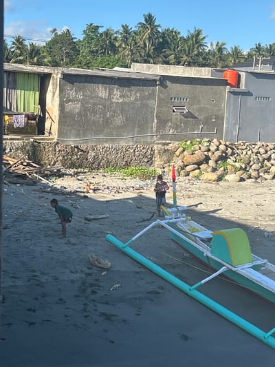 In A R A, Indonesia, a bright, sunny day reveals a beach littered with debris. Two young boys are present on the sandy shore. One boy, wearing a dark green t-shirt and shorts, is crouched near the water. The other boy, in a striped t-shirt and shorts, stands by a colorful outrigger canoe. The canoe is painted green, yellow, and white, with a red and white flag atop a pole. In the background, a concrete building with a corrugated metal roof stands, and beyond that, a dense line of palm trees under a blue sky with scattered clouds. A large orange water tank is visible on the roof of the building. The beach area also features a pile of large rocks and more scattered debris.