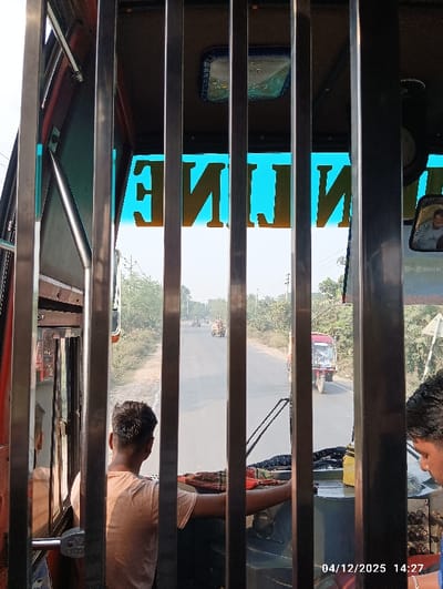 The image displays an interior perspective from a vehicle, likely a bus, looking forward through a partition of six vertical metal bars onto a road in Kaliganj, Bangladesh. In the foreground, a male, identified as the driver, is seen from behind, wearing a light-colored t-shirt. To his right, another male's head and right shoulder are partially visible. Above the windshield, an inverted golden text, which appears to spell "LINE", is illuminated against a bright blue background. A side-view mirror in the upper right reflects a portion of a person's face. A timestamp "04/12/2025 14:27" is present in the bottom right corner.
Beyond the windshield, a two-lane asphalt road stretches into the distance under a hazy, light blue sky. A red three-wheeled auto-rickshaw is observed on the right side of the road, moving away from the viewer. Further down the road, two additional vehicles are discernible. The road is bordered on both sides by unpaved shoulders and dense green vegetation, including trees and bushes.