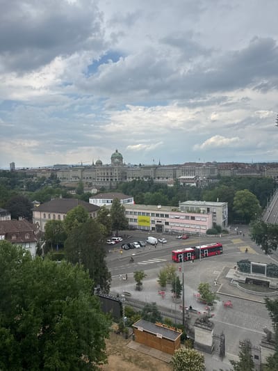 This is an overhead, wide-angle shot of a city street and buildings, captured on a cloudy day. The image is oriented vertically, with the top of the frame showing a sky filled with dramatic, grey clouds, hinting at potential rain or a storm.
The scene depicts a bustling urban environment. A bright red double-decker bus is a prominent object, driving on the paved road. Several cars are also visible on the street, some parked and others in motion. A few people can be seen walking on the sidewalks and crossing the street, some of whom are obscured by the angle.
In the background, a cityscape unfolds with numerous buildings of varying architectural styles. Some appear to be older, grand structures with domed roofs, possibly government or historical buildings, while others are more modern, multi-story edifices. Lush green trees are scattered throughout the scene, interspersed with buildings and lining some of the streets, suggesting a city with ample green spaces.
One building, colored pink, has "PH Bern" written on its side. Another building has yellow signage with text that is partially legible, appearing to be "Grünland ...".
The overall atmosphere is that of a normal day in a European city, with the overcast sky suggesting either early morning, late afternoon, or a generally cloudy day. No specific event seems to be taking place, but the presence of the bus and cars indicates ongoing daily activity.
