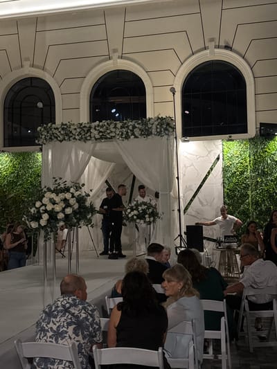 The image depicts an evening event, likely a wedding ceremony, in Kiryat Ekron, Israel. The scene is set in an outdoor or semi-outdoor venue, characterized by a light-colored building facade with three arched dark windows and linear architectural details in the background. Vertical green walls flank the central area on both sides.

A white, chuppah-style canopy, adorned with a dense arrangement of white roses and green foliage along its front edge, is erected on an elevated white platform. Two large floral arrangements, consistent with the canopy's decor, sit on clear pedestals on either side of the platform's approach. Under the canopy, four men are present: one, dressed in a black short-sleeved shirt and dark pants, operates a professional video camera; a second man in a black short-sleeved shirt stands beside him; and two others, dressed in white long-sleeved garments, are positioned further back.

To the right of the canopy, a DJ in a white short-sleeved t-shirt stands behind a console that displays "Zviko Brand." The console holds audio equipment, including a black speaker and a mixer. A tall black stand with a light source is positioned near the DJ setup. A black microphone stand is also visible near the canopy.

In the foreground, multiple guests are seated in white chairs, facing the central ceremony area. Visible guests include a man in a blue and white floral shirt (back to camera), a woman in a black sleeveless top (back to camera), a woman with blonde hair in a light blue top, and an older man in a light-colored collared shirt with glasses. To the far left, a woman with long hair, wearing a brown top and blue jeans, stands looking at a mobile phone. The entire scene is illuminated by artificial lighting, indicating an evening setting.