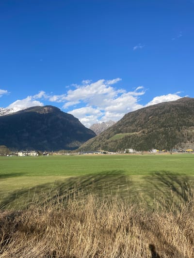 The image captures a landscape in Sterzing-Vipiteno, Italy, oriented vertically. The sky is blue with scattered white clouds. Adjacent to the sky is a mountain range. The lower slopes of the mountain lead to a green field, separated from dry, brown vegetation in the foreground. Buildings are situated at the base of the mountain. Tree shadows are visible on the green field.