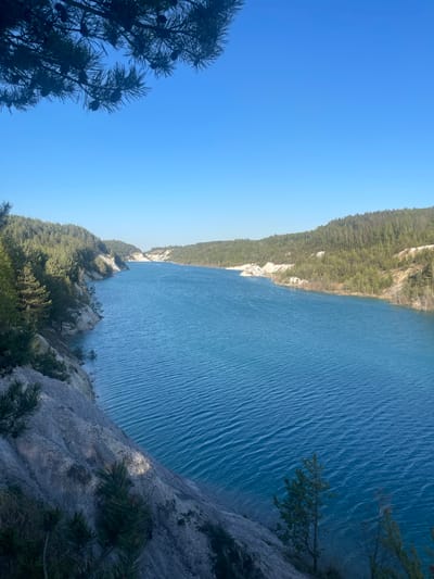 A high-angle, rotated view depicts a blue lake bordered by forested hills under a clear blue sky. The lake surface shows subtle ripples. Trees and rocky outcrops frame the bottom right and bottom left corners, creating a sense of height and perspective. The image captures a natural landscape. The location is within Краснасельскі сельскі Савет, Belarus.