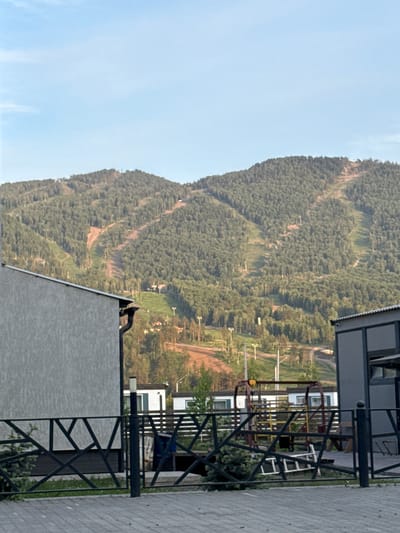 The image depicts a mountainous landscape in Ozernoe, Russia, with forested slopes and visible ski runs. The sky is clear light blue with minimal cloud cover.

In the background, a large, densely forested mountain range extends across the upper half of the frame. Multiple deforested clearings, indicative of ski slopes or runs, traverse the mountain diagonally, appearing as lighter brown or green strips against the darker green forest. Numerous ski lift towers, primarily T-bar or chairlift structures, are visible along these slopes and at lower elevations. Small, scattered structures, possibly resort cabins or buildings, are nestled among the trees on the lower mountain.

The foreground features man-made structures. On the left, a light gray building with a textured finish has a dark gutter and downspout visible on its right corner. A dark roofline is present. On the right, a portion of a modern dark gray building with large dark windows is visible.

A dark metal fence with a geometric pattern of interlocking triangles and trapezoids runs across the lower portion of the image. The ground in front of the fence is paved with gray rectangular pavers. Behind the fence, a small evergreen shrub grows on the left. Towards the center-right, a red and yellow metal structure, resembling a swing set or playground frame, stands on unpaved ground. A silver ladder lies horizontally on the ground near this structure. Further back, several white, modular-style buildings with dark roofs and some with dark or green trim are visible among sparse trees and dirt paths, suggesting accommodation units. A dark lamppost with a light-colored top section stands upright behind the fence. No individuals are present in the scene.