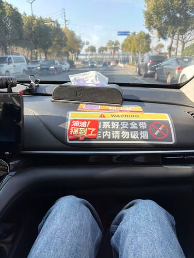 The image presents a front passenger's perspective from within a vehicle, situated in Jianye District, China, facing forward on a multi-lane road under clear daylight. The foreground details the vehicle's black dashboard. On the left, a black electronic device, possibly a dashcam, displays "08:59" and is connected by black cables. Centered on the dashboard is a black tissue box holder bearing the text "滴滴快车" (Didi Kuaiche), with a white tissue partially extracted. Below the tissue box, a rectangular warning sign is affixed. This sign features a yellow banner with "WARNING" and Chinese text, and its main body, in white, displays Chinese characters translating to "Please fasten your seatbelt" and "Please do not smoke inside the vehicle," accompanied by a universal "No Smoking" symbol. A red, irregular-shaped sticker inscribed with "滴滴福利到了" (Didi Benefits Have Arrived) is attached to the left of this sign. Below the dashboard, the passenger's knees, clad in light blue denim jeans, are visible. The midground and background show a paved road with multiple vehicles in motion and parked along the sides. Deciduous trees with sparse foliage line both sides of the roadway, and a pedestrian overpass is discernible in the distant background under a bright, clear sky.