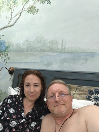 The image shows a couple in bed, possibly at a hotel in Pozzuoli, Italy. The woman on the left is wearing a black and white pajama top with Betty Boop prints. She has medium-length, dark brown hair and is looking at the camera with a slight smile. The man on the right is partially visible, wearing glasses and looking at the camera. He is shirtless, with a black cord necklace. The background features a mural of a landscape with trees, water, and a cloudy sky. A decorative headboard with a floral design is visible behind them. The overall impression is a casual, intimate moment captured in a bedroom.