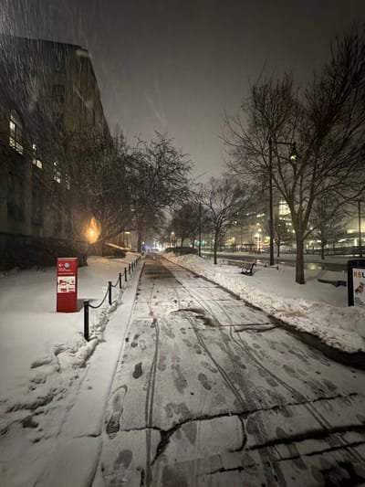 A snowy city sidewalk stretches into the distance. Bare trees line the path, illuminated by streetlights. Footprints and tire tracks mark the snow-covered pavement. A red emergency call box stands on the left. A building is visible in the background. The sky is overcast, and snow is falling. The photo is rotated ninety degrees clockwise.