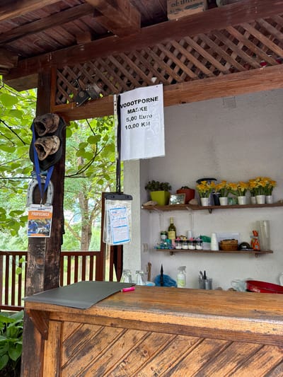 This media file captures a scene from an outdoor setting, possibly a rustic eatery or a covered area with wooden structures and lattice work. The main subjects are various objects displayed on shelves and the ground. On the shelves, there are decorative yellow flowers in white pots, bottles of liquid (including one that appears to be wine), and several small containers that might hold spices or toiletries. A red Coca-Cola branded tin is also visible.
On a lower level, near the wooden beams, are two horse hooves, possibly part of a decorative element or historical artifact. A blue strap and a metal horseshoe are also present. A sign with text in a foreign language is attached to the wooden structure, indicating prices and possibly the type of item being sold: "VODOTPORNE MASIKE 5,00 Euro 10,00 KM". Another visible item is a small, clear plastic pouch containing documents or information, with some blue lettering visible. A brochure or card with the text "PROPUSNICA EUROPEAN RAFTING CHAMPIONSHIP TU rafting N" is also displayed.
The overall setting appears to be outdoors, with lush green foliage visible in the background, suggesting a natural or garden-like environment. The lighting suggests it could be daytime, with diffused light indicating it might be overcast or shaded. There are no people visible in the frame, making the focus entirely on the objects and the rustic environment. The location context provided indicates this scene is from Konjic, Bosnia and Herzegovina, which aligns with the foreign language text on the signs. The scene conveys a sense of local culture or a small business catering to visitors.
