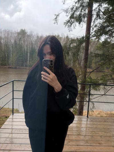 The image is a reflection, likely captured through a window or glass door, showing a person taking a selfie superimposed over an outdoor landscape. The main subject is an individual with long dark hair, wearing a black hooded sweatshirt and dark pants. They are holding a smartphone with a leopard print case in their right hand, positioned to capture their reflection. The person's face is partially visible, with their gaze directed towards the phone screen.
The reflected background reveals a natural environment. A body of water, possibly a lake or a wide river, is visible in the midground, with a light-colored, dry grassy or reedy shoreline. Beyond the water, a dense forest extends towards the horizon under an overcast sky, featuring a mix of coniferous and bare deciduous trees.
In the foreground of the reflected outdoor scene, a wooden deck or balcony with horizontal planks is visible, alongside a black metal railing. This suggests the person is indoors, looking out onto a connected outdoor structure.
The overall atmosphere suggests an overcast day, likely cool or cold given the heavy clothing and the state of the vegetation. The lighting is soft and diffused. A small white patch with an illegible dark logo or text is present on the left sleeve of the hoodie. The location is Mishkino, Russia.