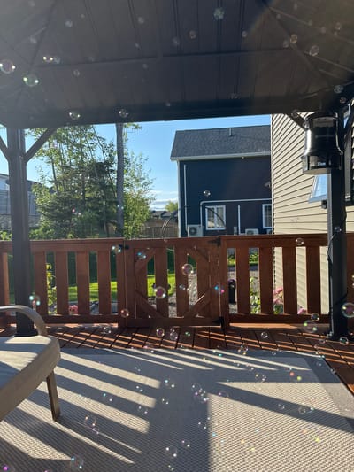 The image depicts a backyard deck area in Dieppe, Canada, under a gazebo. The scene is filled with numerous soap bubbles reflecting sunlight. A grey outdoor chair is positioned on the left side of the frame, situated on a patterned rug. The deck itself is made of wooden planks, and a wooden fence with a gate encloses the area. Beyond the fence, a grassy lawn and trees are visible, along with residential houses under a clear blue sky. A bug zapper device, labeled "DYNATRAP", is mounted on a black pole on the right side of the gazebo. The overall impression is a sunny, outdoor recreational space.