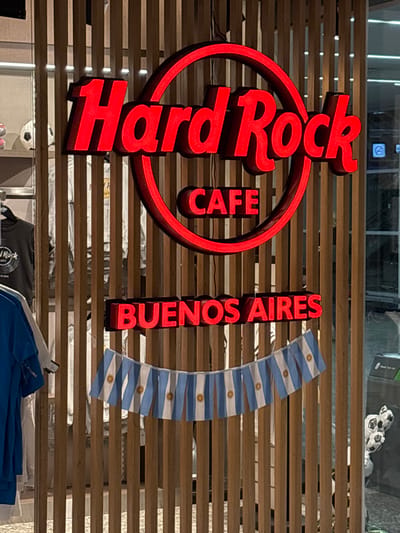 The image shows the exterior of a Hard Rock Cafe in Aeropuerto Internacional Ezeiza, Argentina. The cafe's signage, in bright red neon letters, displays "Hard Rock CAFE BUENOS AIRES". Below the signage, a string of small Argentinian flags hangs across the wooden slat wall. To the left, clothing, including t-shirts with the Hard Rock Cafe logo, is displayed on racks. To the right, a trash receptacle and some decorative soccer ball-shaped balloons are visible. The overall environment appears to be an indoor public space, possibly a commercial area within the airport. There are no people clearly visible in the frame.