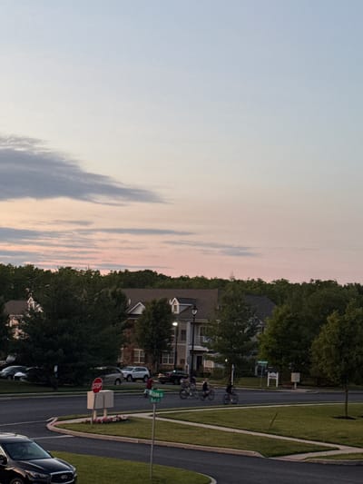 The image captures a suburban street scene in Allen Township, United States of America, during a twilight hour, with a pale blue and pink gradient sky. A line of trees borders the horizon, behind which residential buildings with peaked roofs are visible. In the foreground, a dark-colored sedan is parked on the left side of the frame. A road curves through the scene, with a stop sign positioned on a traffic island in the middle of the intersection. A green street sign indicates "Wilson Ct" and "Rose B." A group of individuals is riding bicycles on the road in the middle ground, heading towards the right of the frame. Several cars are parked in the background along the street.