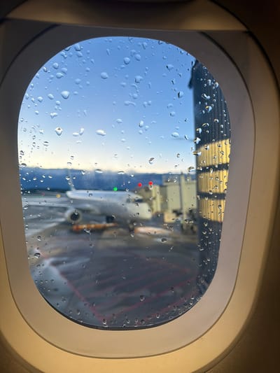 An exterior view is captured from inside an airplane's oval window, which is heavily covered with numerous water droplets, indicating recent rain or significant condensation. The interior window frame is a light beige or off-white hue, illuminated by warm, natural light, suggesting it is daytime.
Through the droplet-obscured glass, the scene outside appears to be an airport environment. In the upper section of the view, a structure with horizontal, possibly reflective, panels is visible, which could be part of a jet bridge or an airport building. Towards the center-right, several distinct colored lights are noticeable: a prominent red light, a green light positioned below it, and two smaller red lights further down. These lights likely belong to ground support equipment, an adjacent aircraft, or airport signage.
The lower-left segment of the window reveals a clearer expanse of bright blue sky, speckled with water droplets. The right side of the exterior view shows a darker, possibly metallic structure, which might be a part of the aircraft's wing or fuselage, or another piece of airport machinery.
The image portrays an airplane parked at an airport gate or on the tarmac, experiencing or having just experienced wet weather conditions. The location context indicates this is likely at Milan Malpensa Airport in Ferno, Italy. No visible text is present in the image.