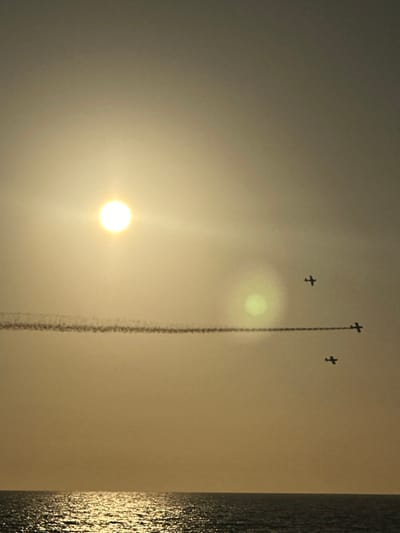 The image depicts an aerial display observed from the coastline of Manhattan Beach, California, United States. The scene is illuminated by the late afternoon or early evening sun, visible as a bright circular orb in the upper-left quadrant of the golden-orange sky, with a notable green circular lens flare to its right. Three single-engine aircraft are silhouetted against the bright sky. One aircraft, positioned horizontally right of center, emits a dense, dark smoke trail extending leftward. A second aircraft flies above and slightly left of the smoke-trailing plane, while a third is positioned below and slightly left, forming a loose aerial formation. The lower quarter of the image reveals the calm surface of the Pacific Ocean, with a shimmering golden path of reflected sunlight extending from the horizon towards the bottom center.