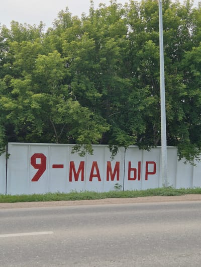 The image depicts a white concrete wall with red Cyrillic lettering that reads "9-МАМЫР". The wall is situated behind a strip of green grass and a gravel embankment, with a paved road in the foreground. Lush green trees with dense foliage form a backdrop to the wall. A tall, slender light pole is positioned to the right of the wall. The sky is overcast and pale. The lettering, "9-МАМЫР", refers to Victory Day, celebrated on May 9th in Kazakhstan, including in the city of Новоишимское. This day commemorates the Soviet Union's victory over Nazi Germany in World War II.