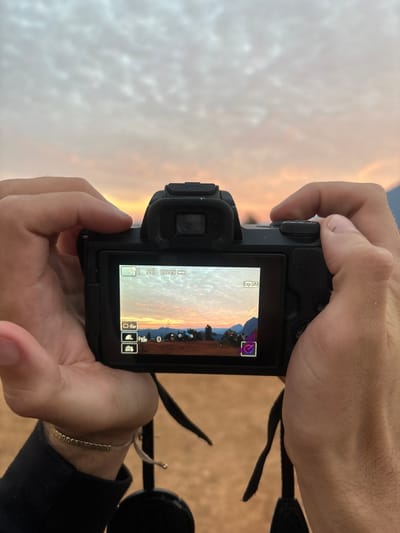A close-up, first-person perspective image depicts two hands holding a black digital camera, possibly a Canon mirrorless or DSLR, at eye level. The camera's rear LCD screen is active, displaying a live view of a scenic landscape. The screen shows a sky filled with mottled, altocumulus clouds displaying hues of orange and pink, indicative of either sunrise or sunset. Below the horizon, a mountainous terrain is visible with several dome-shaped tents pitched on reddish-brown ground, interspersed with sparse trees. Camera interface elements, including "L 59", "02:46", and "Exp. SIM", are visible on the screen. The hands, appearing to be an adult's, grip the camera, with the left wrist adorned by a thin gold-colored bracelet and a string bracelet. A black camera strap and a lens cap hang below the camera. The background above the camera continues the view of the cloudy, colorful sky, matching the scene displayed on the camera's LCD. The setting is an outdoor location with elevated ground, specifically described as Nong Ian, Laos.