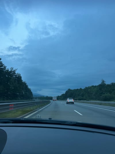 The image depicts a view from inside a vehicle traveling on a highway, likely in Novi Han, Bulgaria. The perspective is from the passenger seat looking forward and slightly to the right. The road is paved with visible white lane markings and a guardrail runs along the right side of the highway.

In the distance, several vehicles are visible on the highway, including a white car in the middle lane and a truck further ahead. The surrounding landscape is dominated by lush green trees that line the highway on the left side. Beyond the trees, hazy mountains are visible in the distance under a cloudy sky.

The sky is overcast with a mix of dark and lighter blue clouds, suggesting either early morning, late evening, or a day with unstable weather. The overall lighting is dim. There are no people clearly visible in the image, only the interiors of cars and the surrounding environment. The scene conveys a sense of travel on an open road under a moody sky. No visible text is discernable.