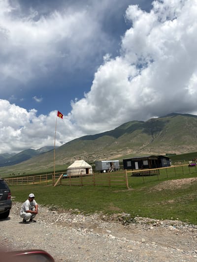 In Semenovka, Kyrgyzstan, a man wearing a white cap, white t-shirt, and grey pants is crouching on a gravelly surface next to a black SUV. In the background, a traditional yurt stands next to several modern structures, including a dark-colored house with a covered porch and a grey trailer-like building. A tall flagpole with a red flag bearing a yellow symbol stands prominently in front of the yurt. The scene is set against a backdrop of rolling, green, mountainous terrain under a partly cloudy sky. A wooden fence encloses the area with the yurt and buildings.