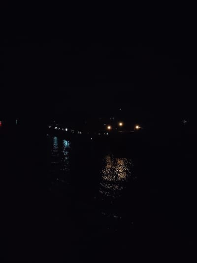 The image depicts a dark, nocturnal scene over a body of water in Pruzhinino, Russia. The vast majority of the frame is deep black, indicating the absence of natural light.
The primary subjects are the water and the reflections of artificial lights on its surface. Distant lights are faintly visible in the background, appearing as small, scattered points. On the left side of the frame, cool-toned lights (blues and whites) are reflected on the water, creating elongated, shimmering patterns. Towards the center-right, warmer, golden-orange lights are reflected, producing a textured, almost liquid-gold appearance on the water's surface. The reflections show subtle ripples and movement in the water. The faint points of light in the distance suggest structures, buildings, or perhaps boats along a distant shore or on the water itself.
No specific activity or event is captured; the image presents a still, contemplative night landscape. The time of day is clearly night. No specific weather conditions are discernible beyond the apparent calm, though the rippled reflections suggest minor water movement. The scene evokes a sense of peace, mystery, or solitude due to the prevailing darkness and scattered light. There is no visible text within the image.