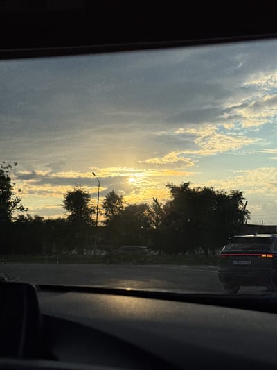 The image depicts a sunset over a rural landscape, likely in Novomar'evskaya, Russia, as suggested by the presence of a vehicle with a Russian license plate. The sun is setting, casting a warm, golden glow on the scattered clouds. The sky transitions from a pale blue at the top to warmer hues of orange and yellow near the horizon. Silhouetted trees and foliage line the right side of the frame, with a streetlamp visible amongst them. In the background, a dark-colored vehicle is parked on the side of what appears to be a road. The license plate on the vehicle is partially visible and reads "0756T8 126". The overall mood is serene and peaceful, characteristic of a quiet evening. The photograph seems to be taken from inside another vehicle, indicated by the dark framing in the foreground.