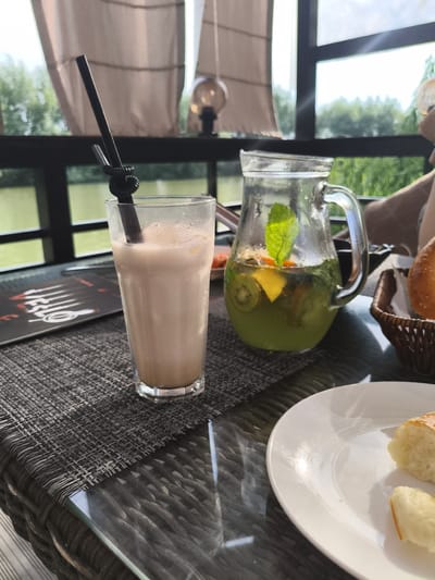 A table setting at an outdoor restaurant in Komsomolskoe, Kyrgyzstan, features a glass of iced beverage with a black straw, a glass pitcher filled with a green liquid, fruit slices, and mint leaves, and a white plate with pieces of bread. The table is covered with a textured placemat and a glass tabletop, overlooking a body of water and trees. A menu is visible on the left, and a woven basket with bread is on the right. The scene is illuminated by natural light, suggesting daytime.