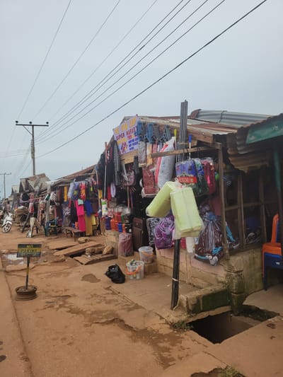 The image depicts a street market scene in Tangu, Nigeria, on an overcast day. The foreground shows a muddy, uneven dirt road with puddles of water. A "NO PARKING BY ORDER" sign stands to the left. The right side of the road features a row of open-air market stalls with corrugated metal roofs and wooden structures. These stalls are laden with various goods, including clothing, bags, plastic containers, and what appear to be household items. Several plastic chairs are visible near one of the stalls. A motorcycle is parked to the far left. In the background, electrical wires are strung across the sky, supported by a utility pole. The overall impression is of a bustling, informal marketplace. There are no visible people in the image.