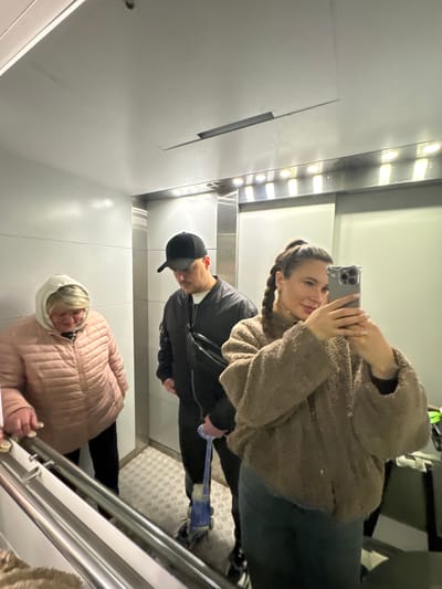 An interior view of an elevator in the city of Gribovo, Russia, shows three individuals. On the left, an older woman with blonde hair, wearing a light-colored hooded top and a pink quilted jacket over dark pants, holds a silver handrail. In the center, a man wearing a black baseball cap, a black bomber jacket, and a black fanny pack across his chest, holds a blue object that appears to be part of a children's scooter or toy. On the right, a younger woman with long brown braided hair and a light brown textured jacket over dark pants holds a silver-cased smartphone, taking a mirror selfie. The elevator features white panel walls, a reflective silver trim with recessed lighting along the ceiling, and a metallic textured floor. A dark piece of luggage is partially visible in the background on the far right.