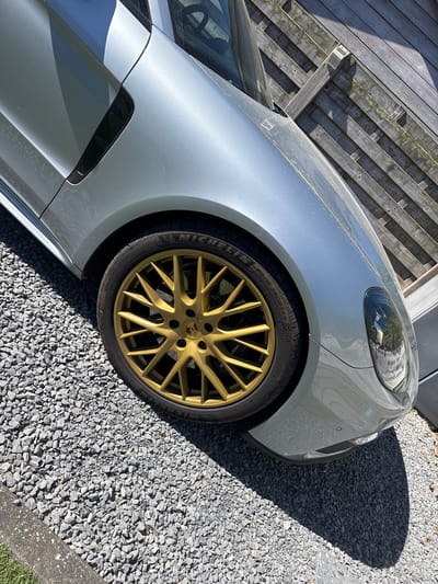 This outdoor shot, taken in Bredene, Belgium, captures the front right quarter of a light grey or silver Porsche, parked on a gravel driveway under bright sunlight. The car features striking golden multi-spoke alloy wheels, with a Porsche emblem visible on the center cap. The tire, a Michelin Pilot Sport 4 S with a "N0" (Porsche-specific) marking, displays text including "275/35 ZR 21" indicating its size.
The car's body appears slightly dusty. Strong shadows are cast on the gravel, indicating a clear, sunny day, possibly morning or late afternoon given the angle of the light. Behind the car, a weathered wooden fence with horizontal slats is visible, partially in shadow. Reflections are seen in the car's headlight, with some indistinct text that might be Dutch or Flemish, given the location. Another distinct reflection of a building structure is also present in the headlight. The image is taken from a low angle, emphasizing the car's wheel and fender.