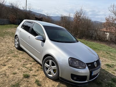 The scene, located in the city of Kastel, Bulgaria, depicts a silver five-door Volkswagen Golf GTI (Mk5) stationary outdoors on uneven ground comprising dry earth and sparse green grass. The vehicle is positioned at an angle, with its front facing slightly towards the left. Key features of the car include darkened headlights, a black lower lip spoiler on the front bumper, a red trim accent above the front Bulgarian license plate 'EB 8303 HB', five-spoke alloy wheels with visibly yellow brake calipers, and tinted windows, including a dark strip across the top of the windshield. The rear driver's side body panel displays multiple circular paint blemishes or marks. The immediate background features dormant deciduous trees, a concrete wall, a metal gate, and a utility pole with power lines. Further back, a white building with an orange-tiled roof is partially visible. Distant mountains appear under a partly cloudy sky. No individuals, actions, or interactions are present in the image.