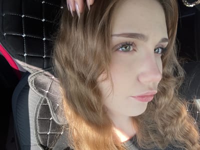 The image shows a close-up, profile view of a young woman's face. She has long, wavy, light brown hair. Her eyes are green with visible eyelashes and eyebrows. She is wearing subtle makeup. Her lips are slightly parted and appear to be a natural pink color. The background consists of a dark, quilted material, possibly part of a car seat, with some sunlight casting shadows. There is also a hint of red fabric visible. The context of Новая Отрадовка, Russia, is not discernible from the visual information provided in the image.