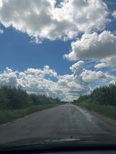 The image captures a road through a rural landscape in Gubachevo, Russia. The road is asphalt and appears wet, with several puddles reflecting the sky. Lush green trees and vegetation line both sides of the road, forming a dense border. The sky above is a vibrant blue, populated by numerous fluffy white cumulus clouds, some of which are quite large and dramatic. The perspective suggests the image was taken from a vehicle moving along the road, as the foreground shows the dark dashboard of a car. The road stretches into the distance, disappearing over a slight incline. The overall scene is one of a pleasant, possibly post-rain, summer day in a natural setting.