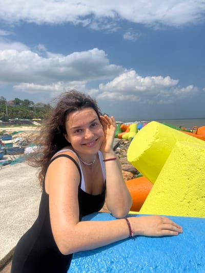 A young woman is captured in a close-up, outdoor portrait against a backdrop of the sea and sky in Phú Thủy, Vietnam. She is positioned to the left of the frame, leaning on a bright blue, textured barrier. Her dark, wavy hair is gently blowing to the left. She is wearing a black tank top with a white strap visible. A white beaded necklace adorns her neck. Her right hand is raised, with fingers lightly touching her cheek, and a dark red bracelet is visible on her wrist. Her left arm rests on the blue barrier, and her hand is open with fingers slightly extended.
In the background, the ocean stretches out under a bright blue sky dotted with white, fluffy clouds. A row of vibrantly colored, large concrete tetrapods, in shades of yellow, orange, and blue, form a breakwater along the coastline. To the left of the tetrapods, a beach area is visible with scattered fishing nets and small boats. Further in the distance, lush green trees and buildings are discernible, suggesting a coastal town or village. The lighting indicates it is daytime, with the sun casting shadows and highlighting the textures of the objects.