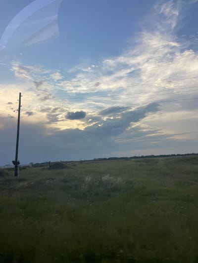 The image depicts a wide, open grassy field under a sky with scattered clouds. A solitary utility pole stands on the left side of the frame, with power lines stretching horizontally across the upper portion of the image. In the distance, a line of trees or structures can be seen along the horizon. The lighting suggests it is either early morning or late evening. The scene is devoid of people or any discernible activity. The environment appears to be rural or semi-rural, possibly on the outskirts of Novoyeniseyka, Russia, given the characteristic flat terrain and the presence of utility infrastructure.