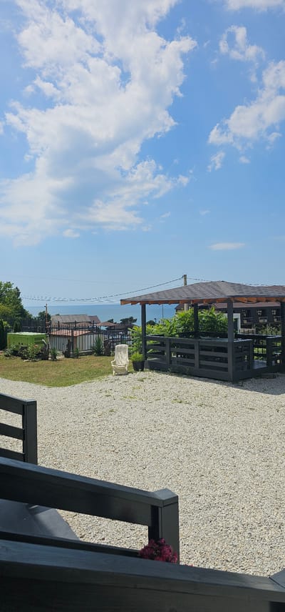 The image depicts an outdoor scene on a sunny day, with a blue sky filled with scattered white clouds. In the foreground, a dark-colored railing and stairs lead down to a gravel pathway. To the right of the pathway is a dark wooden gazebo with a sloped roof, housing tables and chairs. Beyond the gazebo, a light-colored plastic chair sits on a patch of grass. In the mid-ground, there is a green generator, some potted plants, and a black metal fence. Further in the distance, a body of water, likely the Black Sea, is visible, with several buildings and structures along the coastline. The overall setting appears to be a seaside resort or property in Lermontovo, Russia, offering a view of the sea.