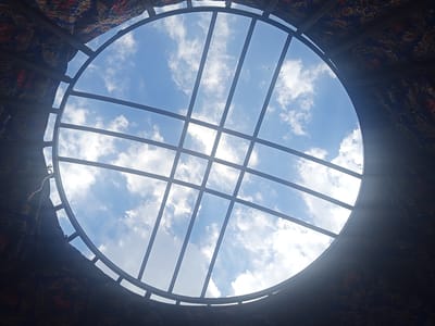 The image shows a view looking up from inside a yurt towards the sky through a circular opening in the roof. The opening is framed by a metal lattice structure and surrounded by the decorative fabric of the yurt's interior. The sky is blue with scattered white clouds. The yurt appears to be located outdoors, possibly in a rural area given the context of its typical use in Kyrgyzstan. The image captures the dome-shaped ceiling of the yurt and the daylight filtering through.