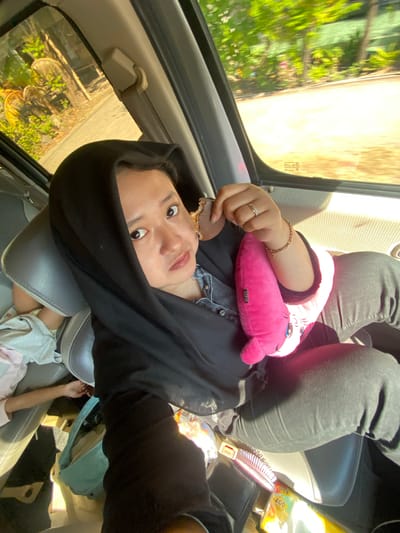 A woman wearing a black hijab and black clothing is seated in the passenger seat of a vehicle. She is holding a small, pink, plush toy in her right arm and a piece of food in her right hand. She is looking towards the camera. A child is partially visible in the backseat. The vehicle appears to be in motion, as indicated by the blurred trees and foliage visible through the windows. The location is identified as Kebondalem, Indonesia.