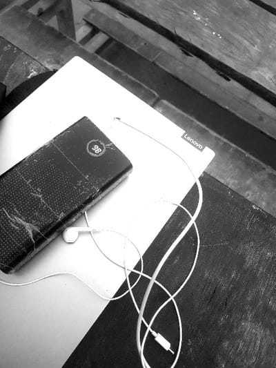 The black and white image displays a still life composition of electronic devices resting on a surface. The shot is taken from a slightly angled overhead perspective.
In the center, a white or light-colored laptop or notebook is partially visible, with the "Lenovo" logo clearly legible on its upper-left corner. Resting on the laptop, towards the bottom of the frame, is a dark-colored power bank. This power bank appears well-used, showing visible scratches and wear on its textured surface. A small digital display on the power bank shows the number "28", likely indicating a battery percentage, possibly with a faint battery icon beside it.
Coiled and spread across the laptop and the adjacent surface are white wired earphones. One earbud is visible towards the bottom-center, and the 3.5mm audio jack is visible towards the upper-right corner of the image.
The objects are placed on a surface that appears to be a wooden table or desk. The surface is divided into two distinct areas. To the left and top of the image, the surface consists of wooden planks, showing visible grain and seams between the planks. To the right and bottom, the surface is a darker, more uniform texture, possibly a different part of the same table or a separate dark mat.
The overall tone of the image is monochromatic, emphasizing textures and shapes. There is no activity taking place; it is a static arrangement of personal items.