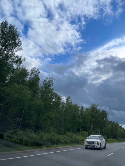 A white car is parked on the shoulder of a paved road in Novyi Byt, Russia. Adjacent to the road is a dense line of trees. The sky above is partly cloudy, with patches of blue visible between the clouds.