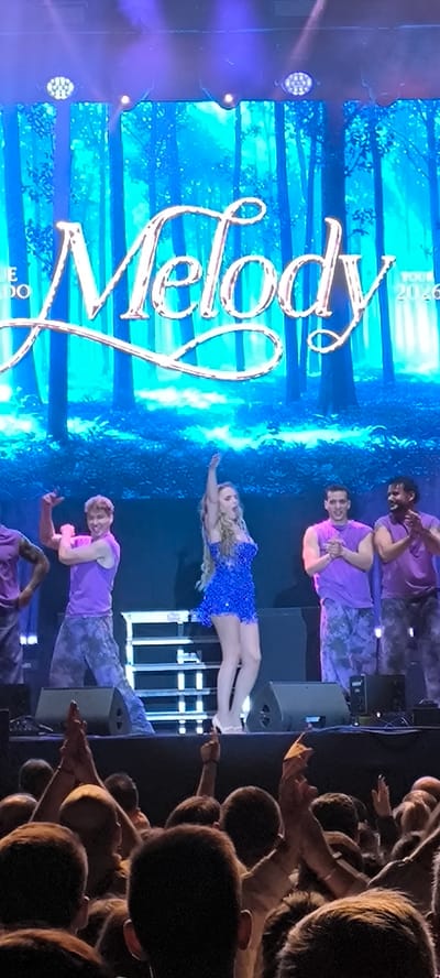 A photograph taken at an outdoor concert in Cerdanyola del Vallès, Spain, depicts a female performer in a blue sequin dress and white high heels on a stage, flanked by four male dancers in purple tank tops and patterned pants. The backdrop features a digitally projected forest scene with the name "Melody" prominently displayed. A crowd of spectators is visible in the foreground, with many raising their hands in applause. Speakers are positioned on the stage floor. The environment suggests an evening or nighttime concert setting with stage lighting illuminating the performers.