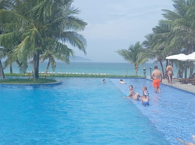 In Dong Ninh Hoa, Vietnam, a large, crystal-clear blue swimming pool is the central focus of this scene. The pool is adjacent to a tropical beach with lush palm trees framing the view. The ocean, a vibrant turquoise, stretches out to the horizon, meeting a hazy, pale blue sky. Several people are enjoying the pool and its surroundings.
In the foreground, the water is a deep, inviting blue, with intricate mosaic tiles visible on the pool floor. Several individuals are in the pool: a woman with dark hair is sitting on the edge, leaning back and looking to her left; a man in orange shorts stands in the shallow end, facing away from the camera; and a young boy in blue swim trunks is playing in the water near the edge. Further in the pool, a couple, a woman in a bikini and a man, are interacting.
On the far side of the pool, a paved area is lined with lounge chairs and large white umbrellas, suggesting a resort or hotel setting. Several people are visible on this paved area, including a woman in a bikini and other figures in the distance. The background reveals a verdant landscape with more palm trees and the distant mountains of Vietnam.