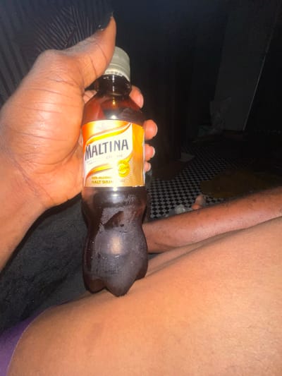 In this close-up, low-light image, a hand holds a sweating bottle of Maltina, a non-alcoholic malt drink, positioned against a person's thigh. The bottle's label, partially visible, reads "MALTINA" and indicates it is "MULTI-VITAMIN ENRICHED" and a "NON-ALCOHOL MALT DRINK". A black and white checkered pattern is visible in the background, suggesting an indoor setting. The overall lighting is dim, with the focus on the bottle and the hand. The water droplets on the bottle suggest it is cold. No specific activity or event is clearly depicted, beyond the act of holding the drink. The image is cropped tightly, revealing only a portion of a person's leg and hand, and the product. The location context of Akowonjo, Nigeria, is provided, but not visually evident in the image itself.